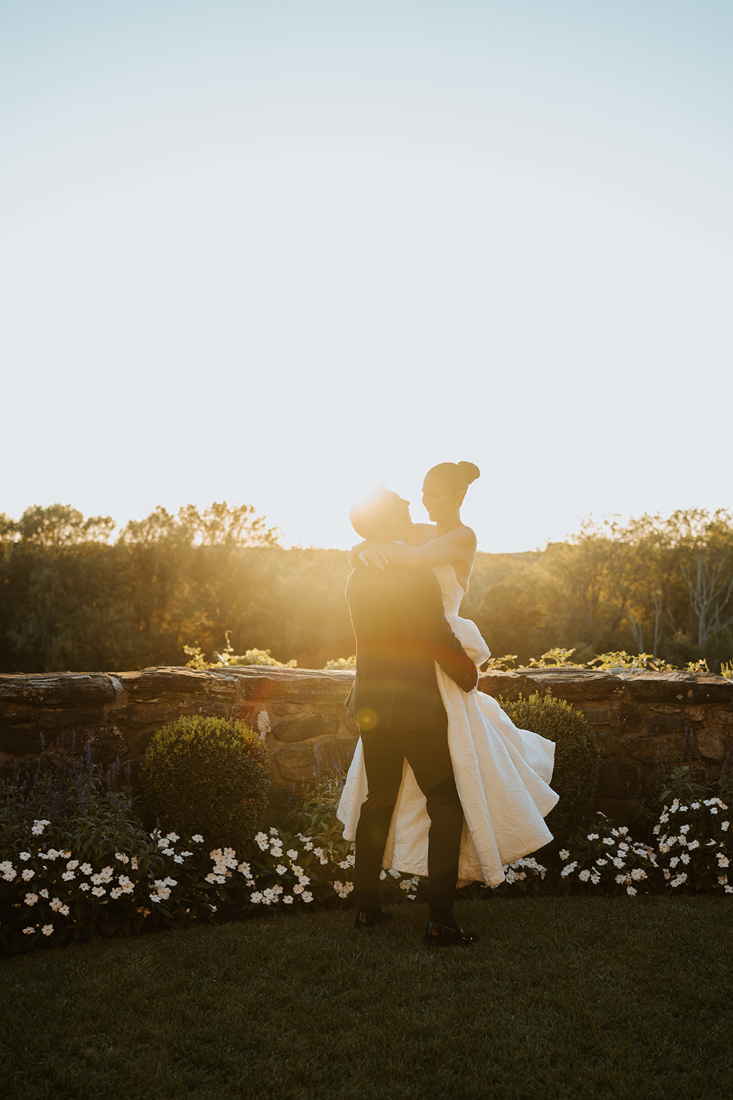 Groom lifting and holding his bride by a stone wall at sunset, bathed in a warm golden hour flare.